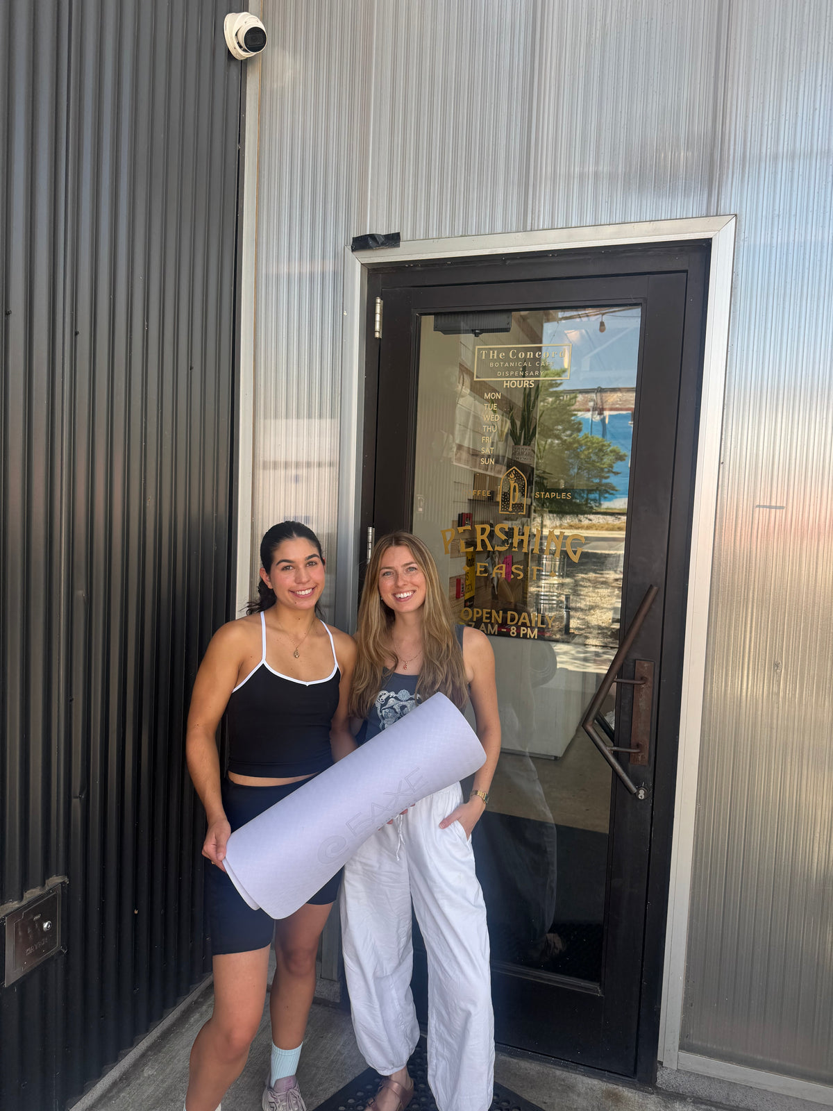Two women standing outside the Pershing cafe doorway holding a light purple yoga mat. One woman is wearing a black tank top and shorts, and the other is wearing a gray tank top and white pants.
